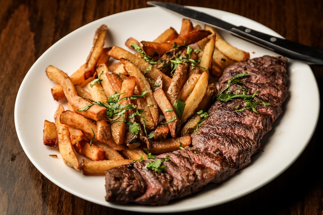 Steak panggang medium di atas piring putih dengan kentang dan taburan peterseli, di meja kayu gelap